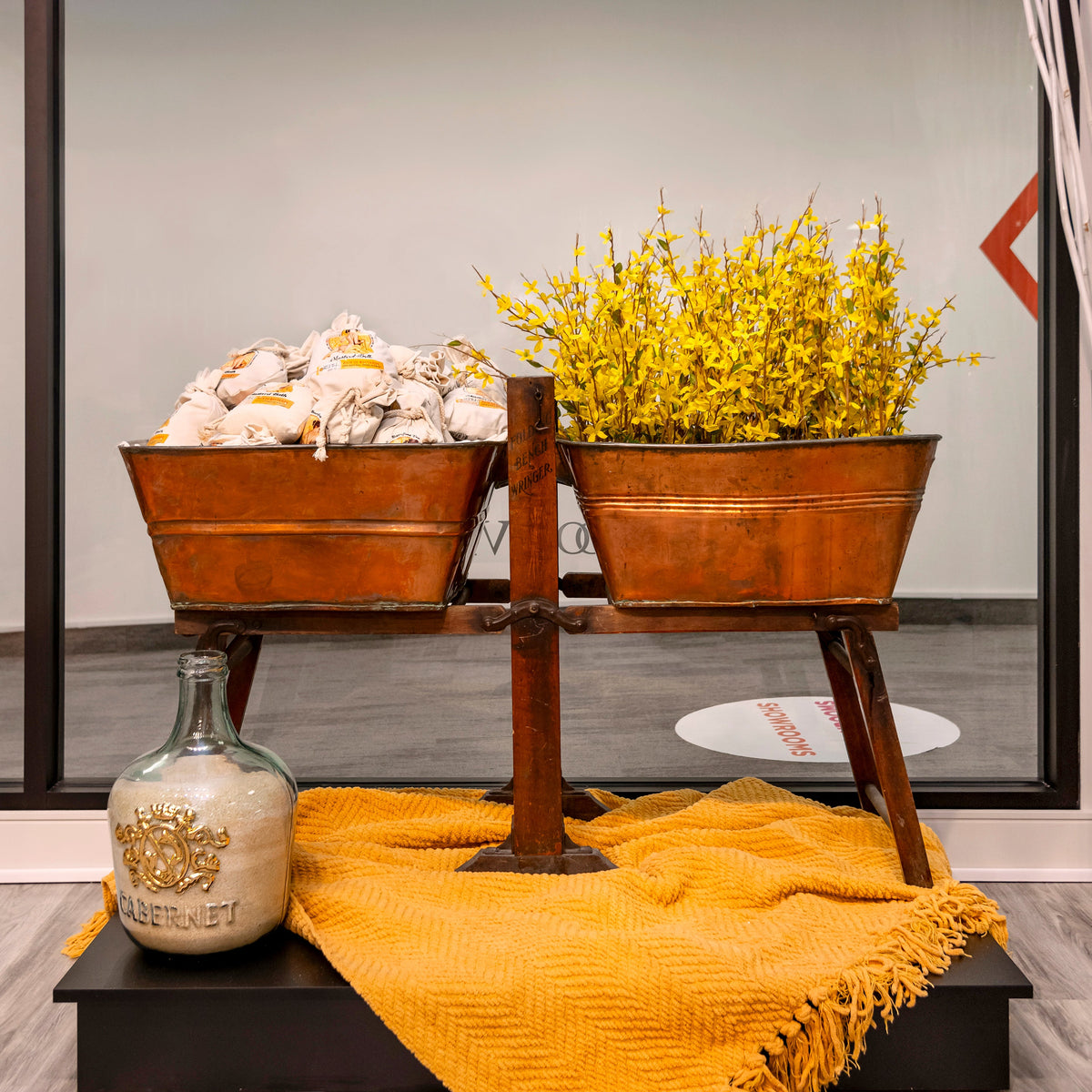 Mustard Bath refill bags in vintage bin displays with a large glass bottle of Mustard Bath Soak, yellow flowers, and a yellow blanket.