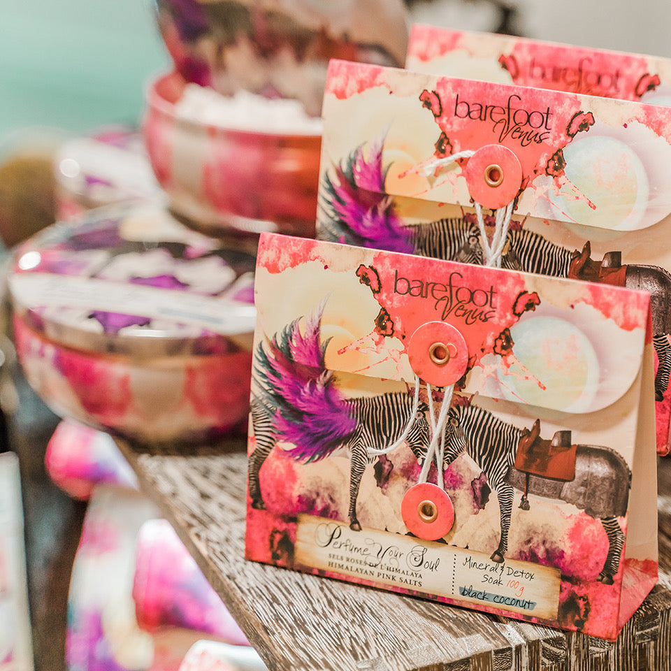 Envelopes of Barefoot Venus Black Coconut Himalayan pink salts displayed with other black coconut products blurred in the background.
